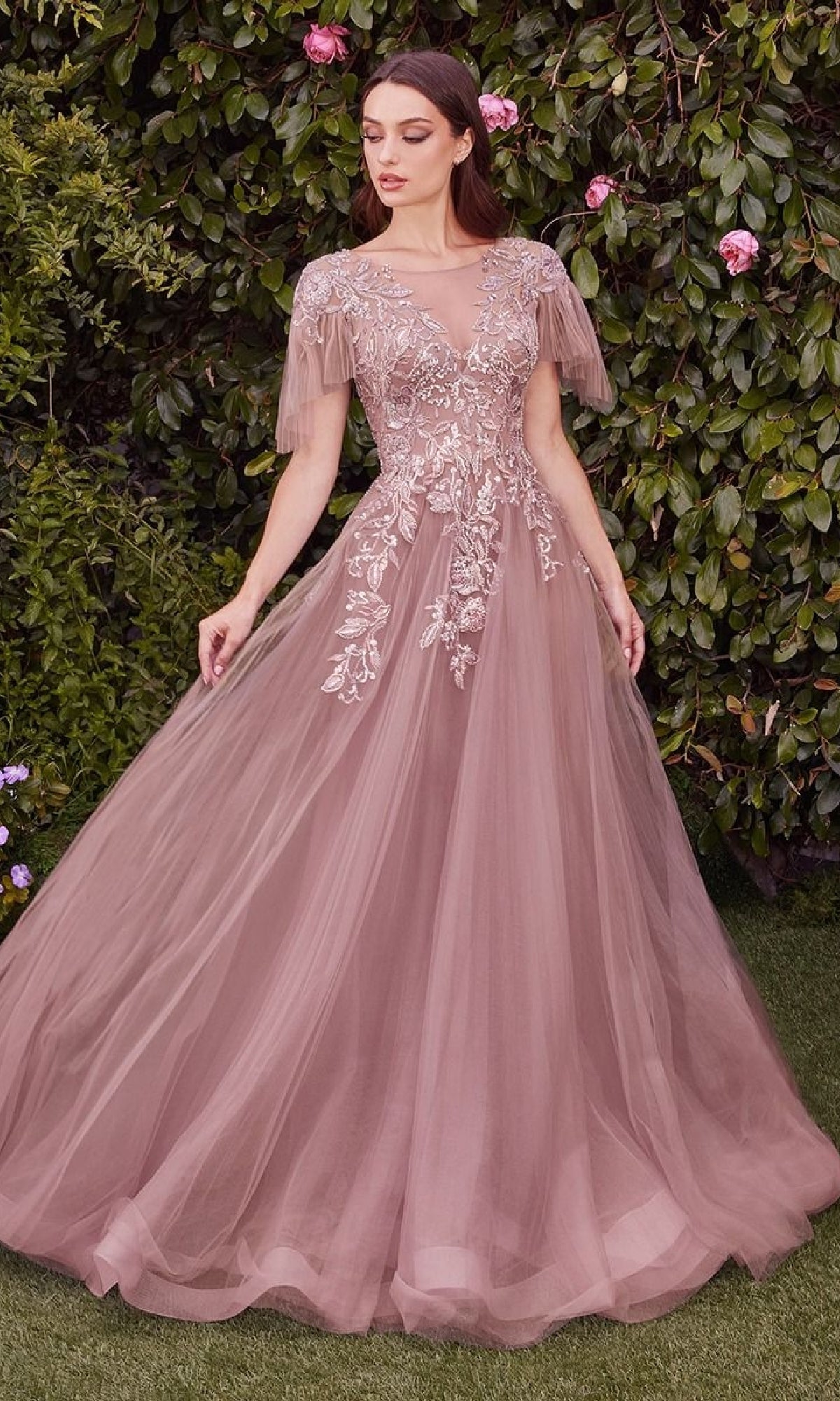A woman stands outdoors in the Andrea & Leo A1351 short-sleeve prom ball gown, a mauve tulle A-line dress with floral and sequin embroidery and sheer flutter sleeves, posing gracefully amidst leafy greenery.